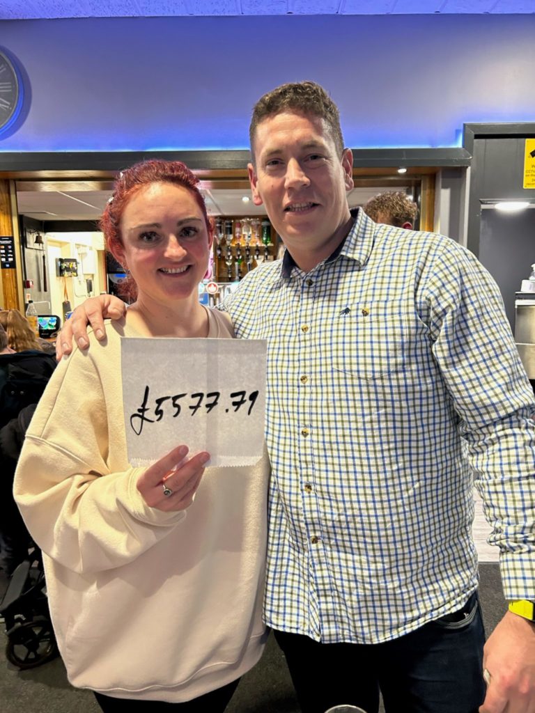Carla Spence is on the left of the image holding a paper displaying the total raised (£5577.79) from ticket sales. On her right is a man with his arm around her shoulder.