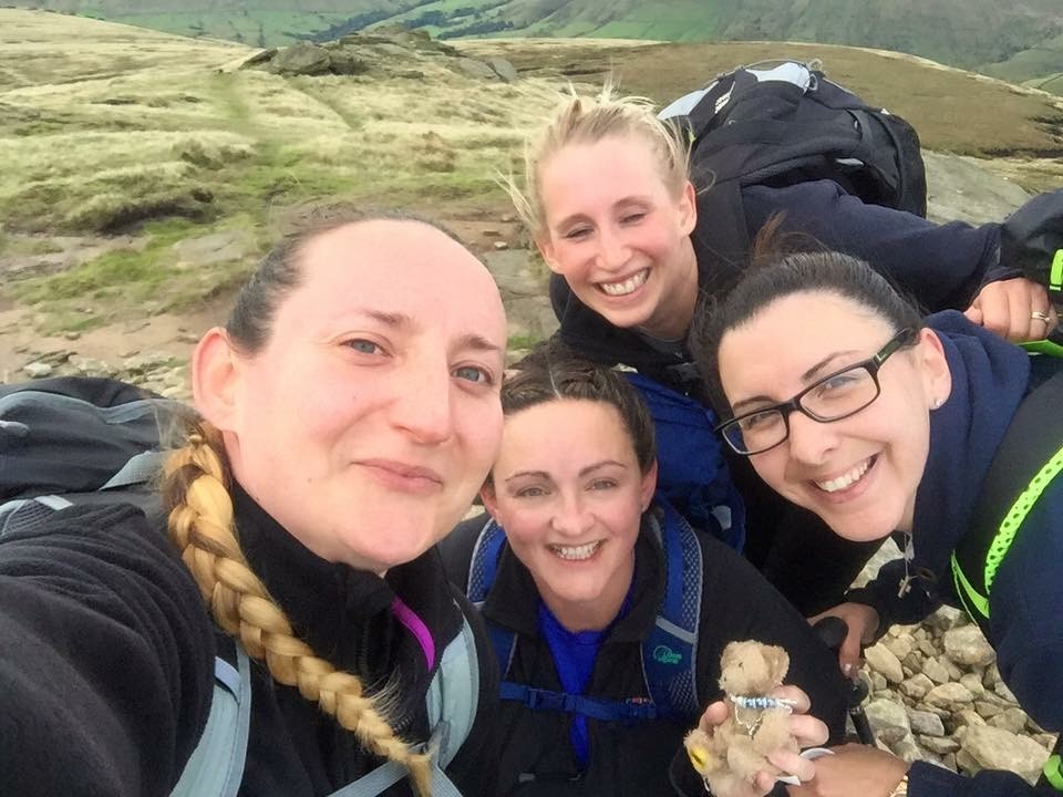 Four women smiling at the top of a grassy hill wearing hiking jackets and backpacks. One of the women is holding a small teddy bear.