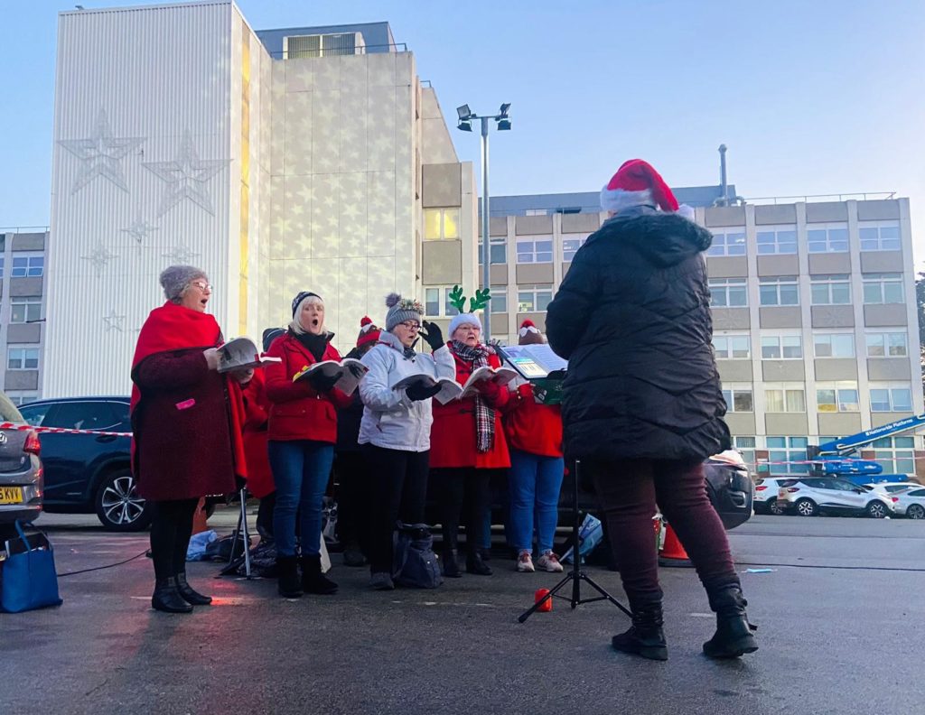 Several individuals dressed in festive-wear such as santa hats and red coats stand in front of the hospital and star illuminations, holding their choir books and singing