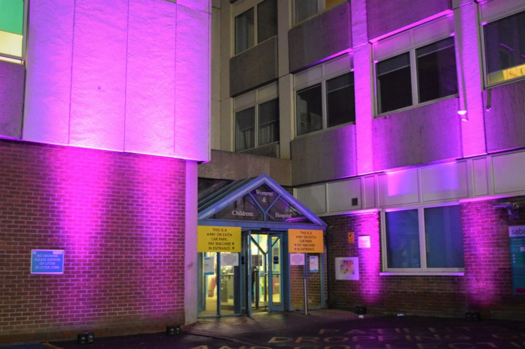 Image description: Image shows the entrance to the Women's and Children's Hospital at night. The doorway is illuminated by two beams of purple light each side.
