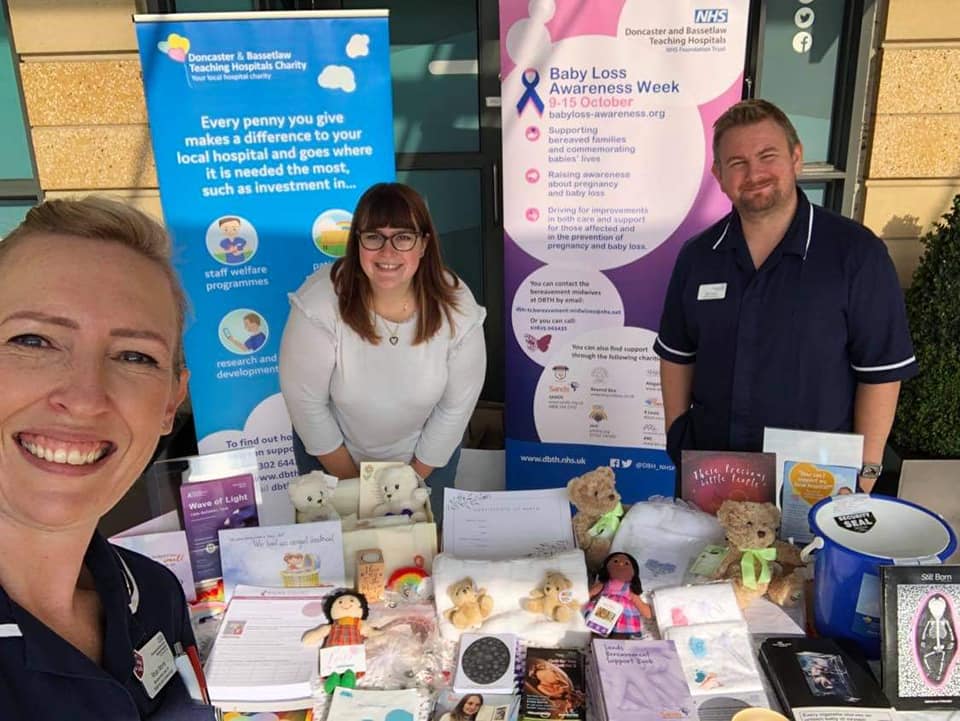 Rhian is taking a selfie with Sarah in the centre and Matt on the right. The awareness stand contains dolls, small teddy bears, a collection bucket and written materials. Behind Sarah and Matt are two banners promoting the charity and baby loss awareness.