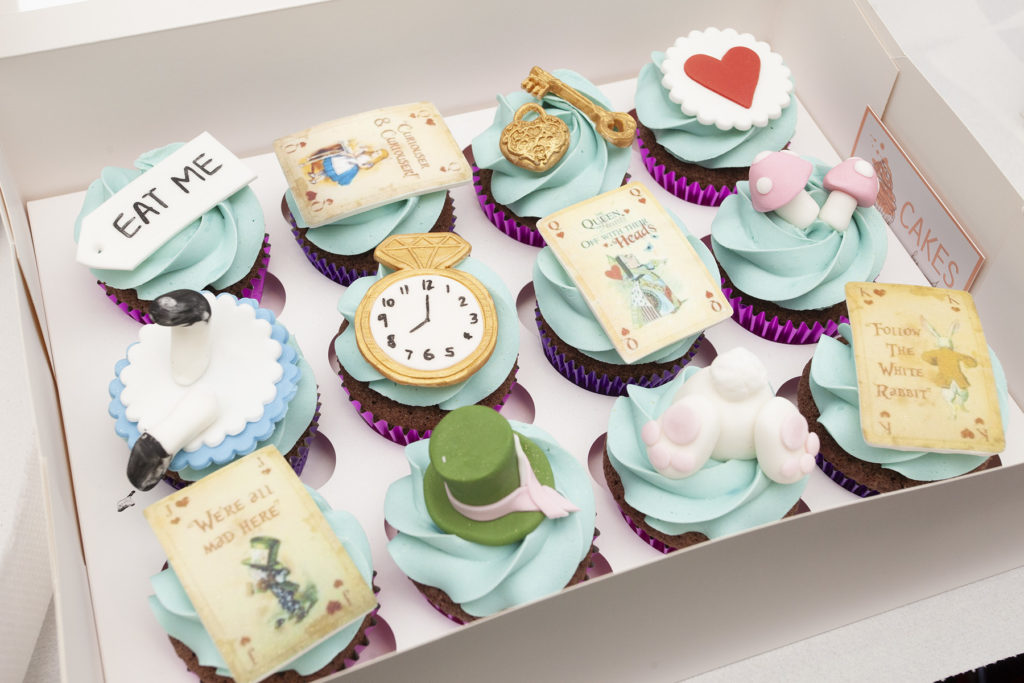 A photograph of Alice in Wonderland themed cupcakes, donated by Cakes by Amy. They had light blue icing with different recognisable symbols of the story, including a pocket watch, play cards, top hat and hearts. 