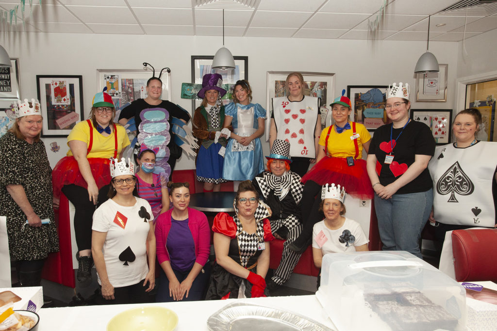 A big group photograph of Paediatric Ward colleagues dressed as Alice in Wonderland characters for World Book Day. Notable characters include Alice, the Mad Hatter, playing cards, Queen of Hearts and the Cheshire Cat. 