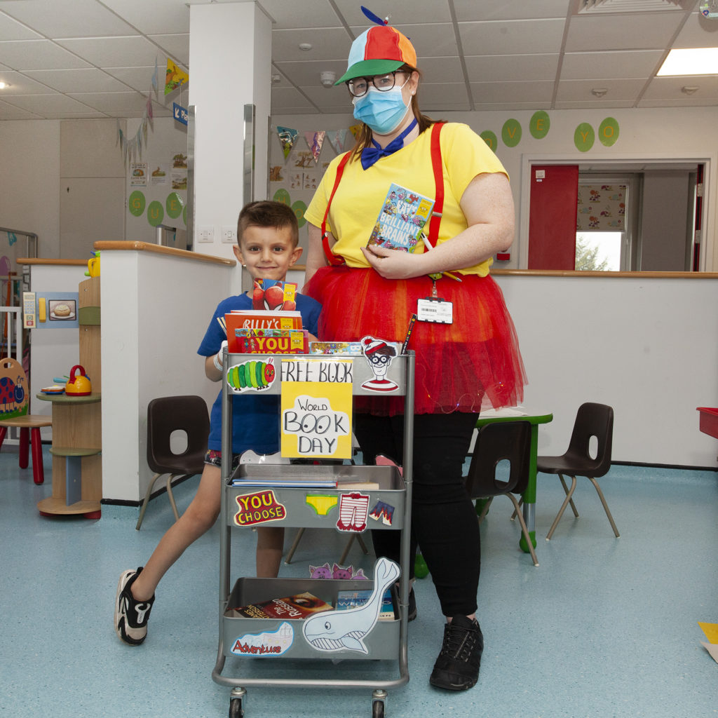 A photograpgh of a young, male patient and staff member dressed as one of The Twins holding a book next to the World Book Day trolley, donated by The National Literacy Trust. 