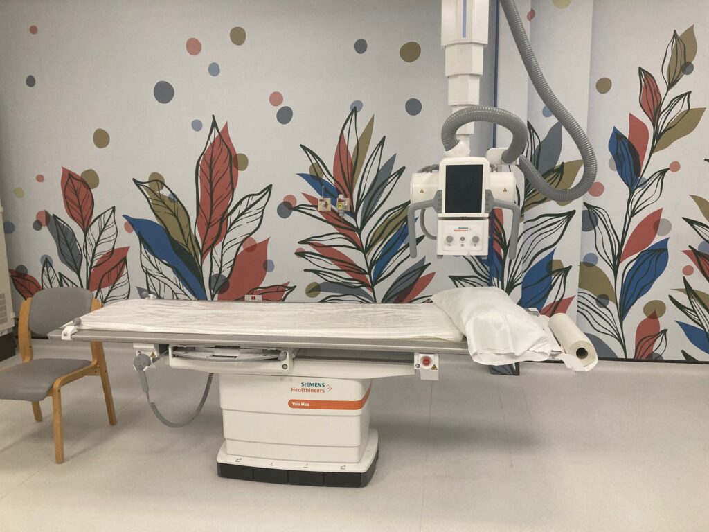 A photograph of x-ray room 4, featuring the new scanning machine and colourful artwork on the walls. The artwork depicts green, orange and blue leaves with a black leaf outline and coloured dots. 