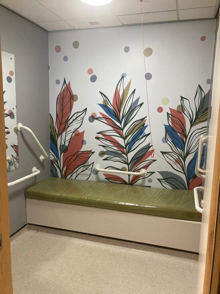 X-ray Room 4 Changing room featuring the new bespoke artwork. Colourful leaves in orange, blue and green are printed across the walls behind a cushioned seat in small changing room space. 