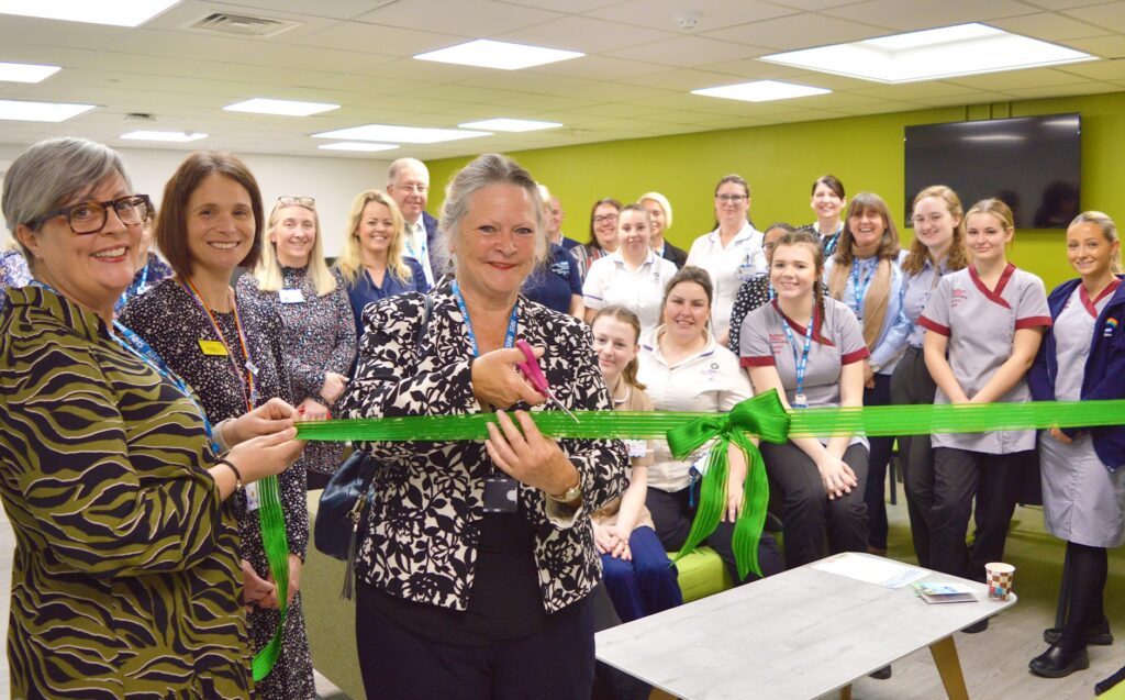 A photograph of the Student Hub opening. Suzy Brain England OBE, Chair of the Board, is in the centre cutting a bright green ribbon. Behind her, Sam Debbage, Director of Education and Research, Zoe Lintin, Chief People Officer and a group of students, and members of the Education Team are smiling at the camera. . 