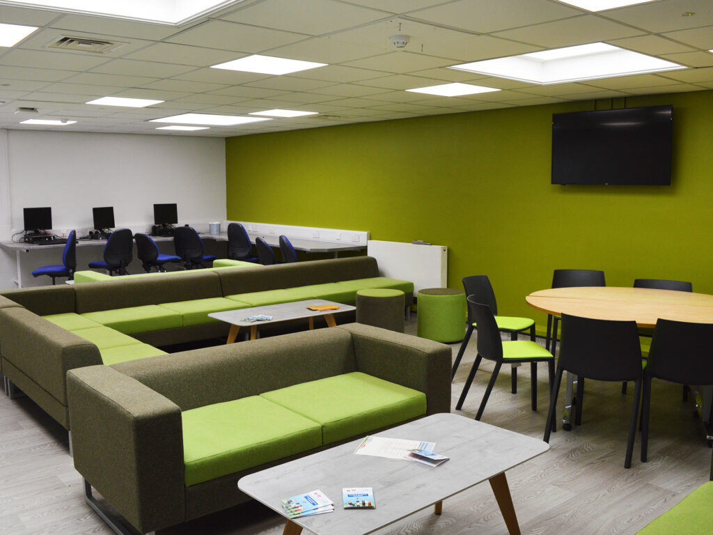 A photograpgh of the Student Hub. The room has one bright green wall and is filled with green sofas and chairs. At the back sits a PC suite and there is a TV on the wall. 