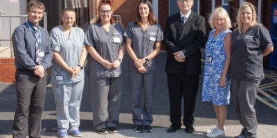 A photograph of all seven members of the AAA Screening Programme Team. They are stood outside in the sunshine in their clinical uniform and office wear. All are white, two are male and five are female staff members.