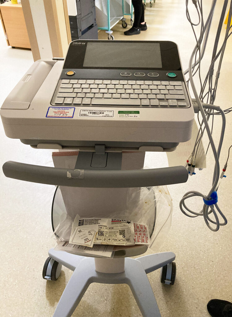 A photograph of the ECG machine donated by the League of Friends. It is mobile with small wheels an attached to a waist-height stand. Long grey cables with detectors attached are plugged into the machine which has a small screen and keyboard.