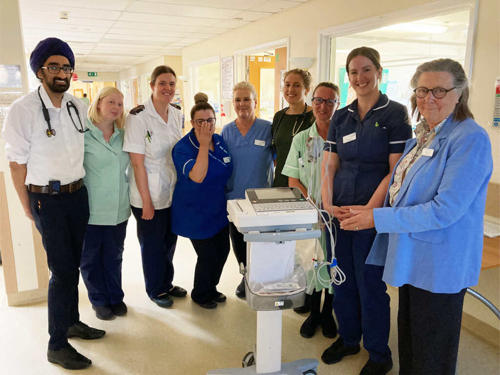 Photograph of clinical colleagues from Ward A4 in uniform with Mrs Philippa Farr, President of the League. They are stood in a semi-circle behind the donated ECG machine.