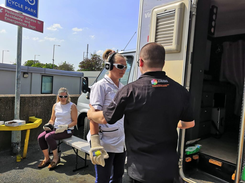 A photograph of hospital staff participating in the Dementia Tour Bus experience. A female staff member in white clinical uniform is stood at the entrance to the bus with a facilitator, wearing special goggles and headphones.