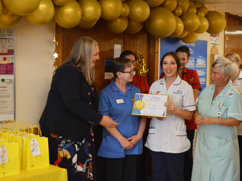 A photograph of a female representative from NHS England, wearing a colourful dress and black blazer, presenting a certificate with a gold medal on it. Ward 17 colleagues are stood to the right receiving the award dressed in their blue and white clinical uniforms. Above their heads hang gold balloons. 