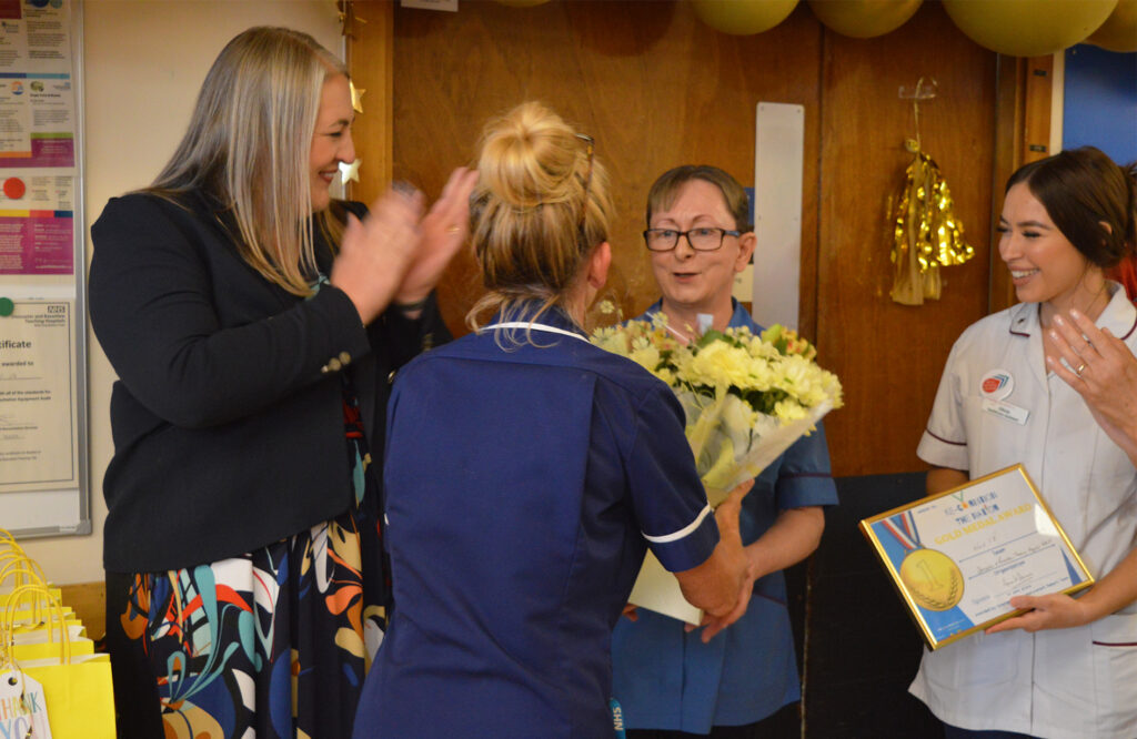 A photograph of Jo McQuade receiving a bouquet of yellow flowers during the award celebrations. Her colleagues surround in applause. 