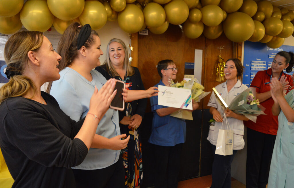 A photograph of Olivia Ada receiving a bouquet of flowers during the award celebrations. Her colleagues surround in applause. 