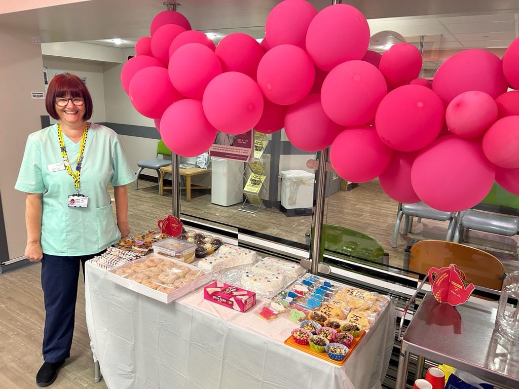 A member of staff is stood by a table filled with cakes, buns and doughnuts. A large collection of pink balloons are pinned above the table.