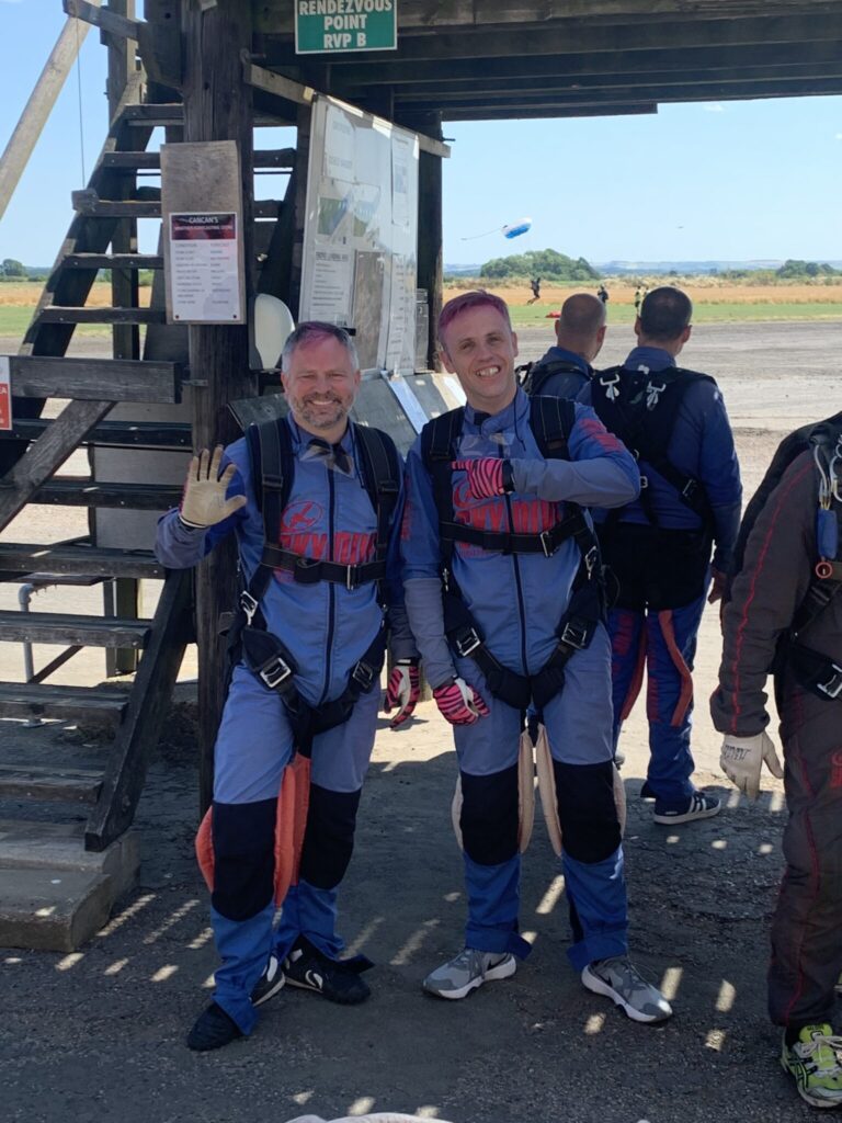 Two men are posing in blue jumpsuits which have the words 'Skydive Hibaldstow' written on them. They are both smiling. One is waving, one is pointing to the other.