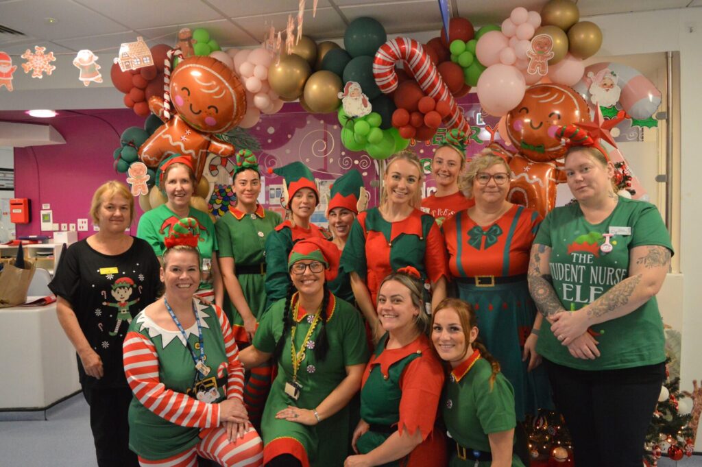 12 Children's Ward staff dressed in green and red elf costumes grouped together smiling in front of the reception desk. 