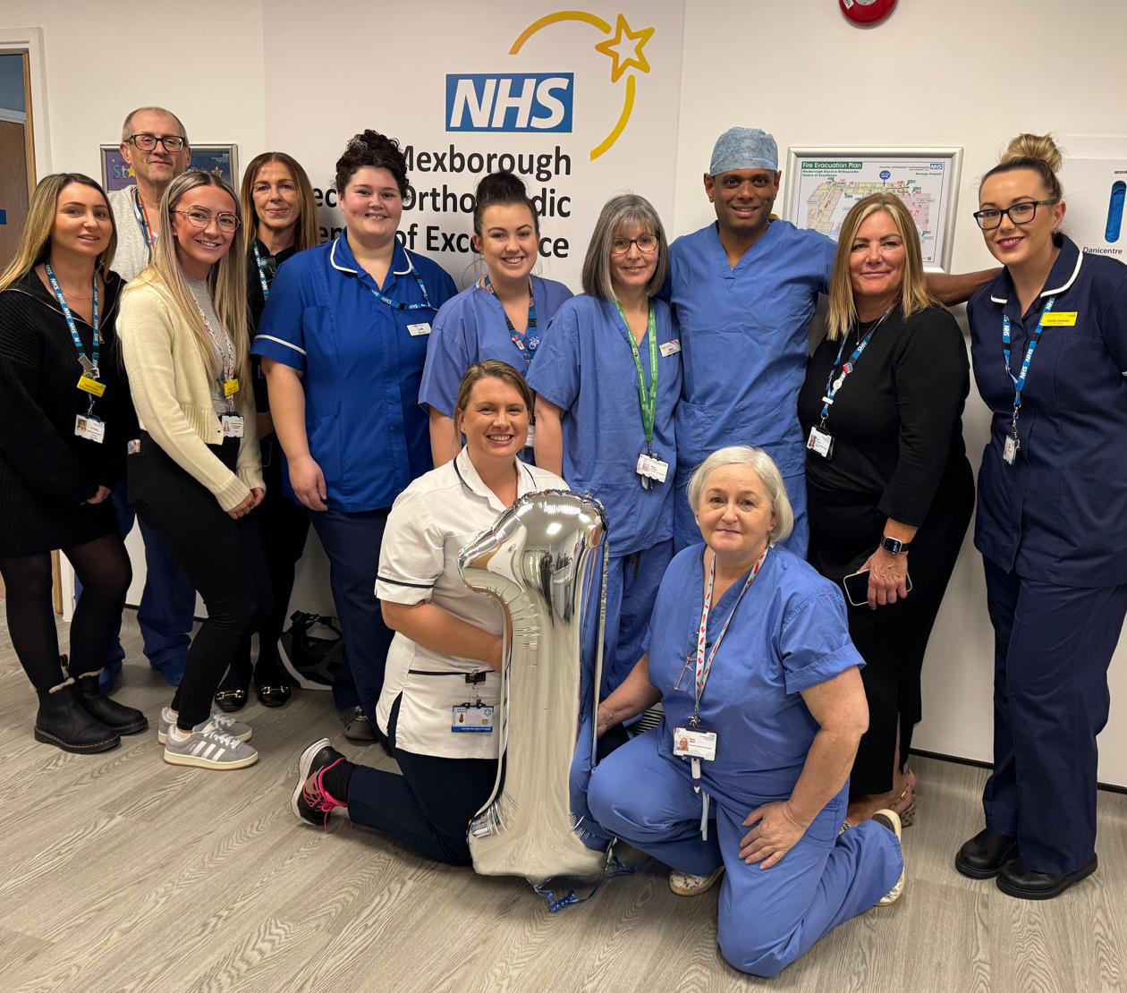 A group photo of 12 staff members in their blue nursing and theatres uniform gathered around a silver 1 balloon