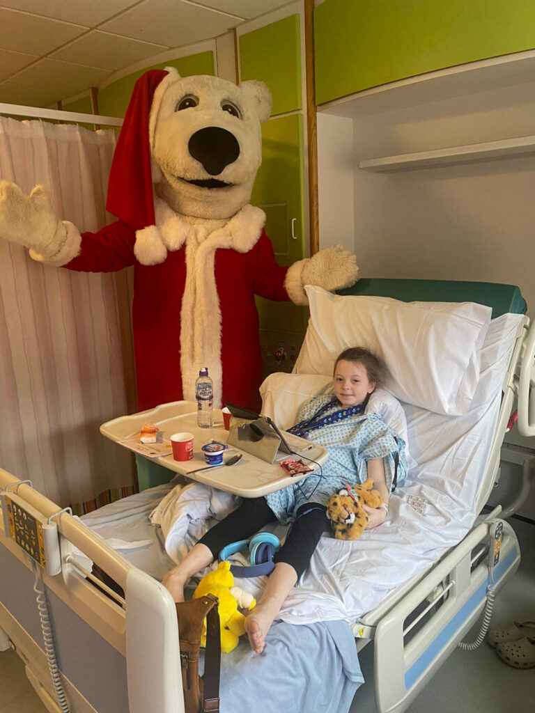 A person in a very tall white polar bear costume and Santa outfit posed next to a young patient in a hospital bed.