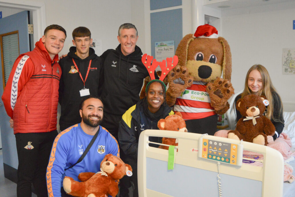 Players are dressed in their black and red branded kit next to a young patient holding a teddy bear in a hospital bed. 