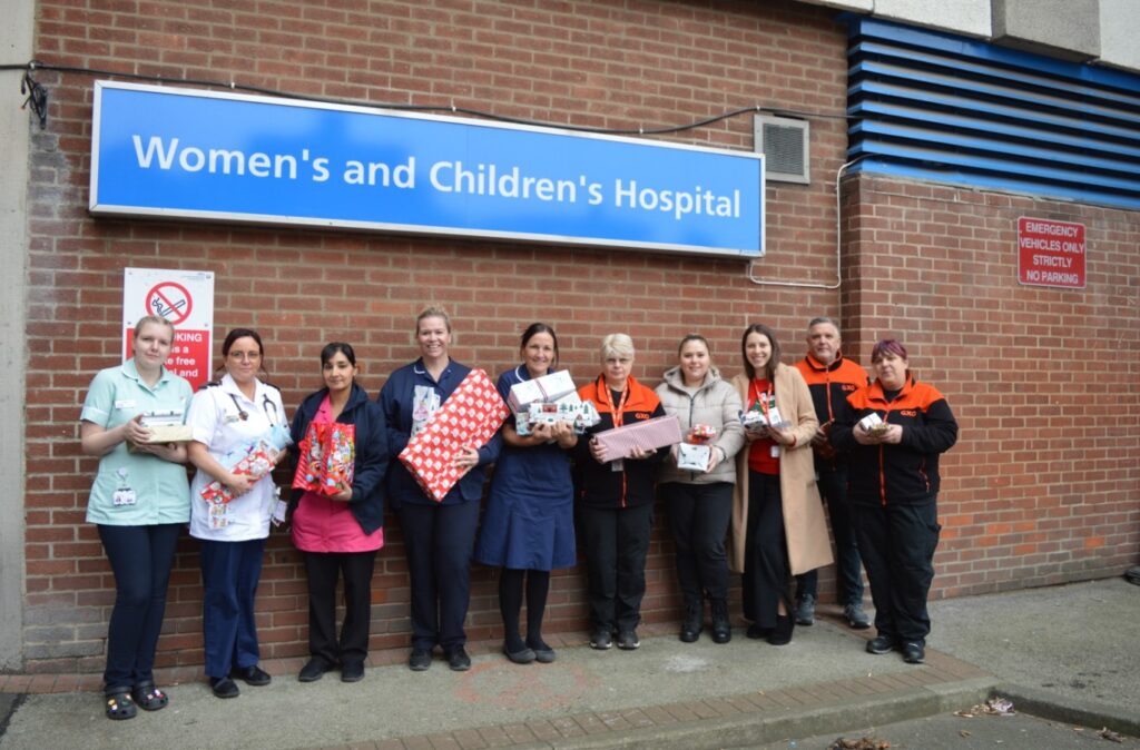 GXO team in orange and black uniform pose holding presents with nursing staff from the Children's Ward underneath the Women's and Children's Hospital sign