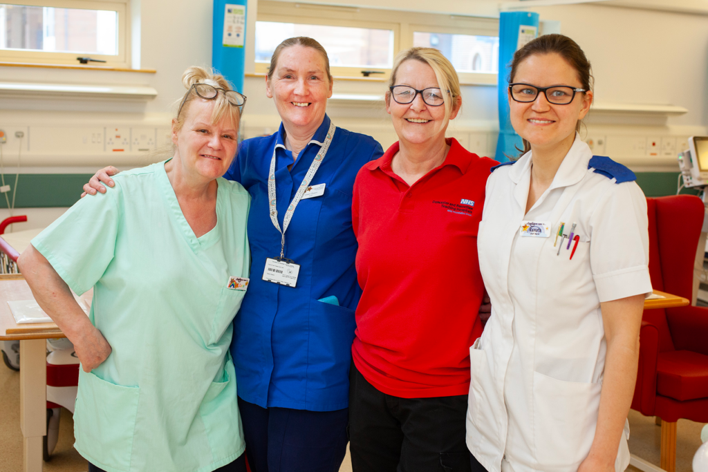 Photograph of four female hospital colleagues smiling at the camera. One is wearing a light green uniform, one a dark blue uniform, one a red shirt and the fourth a white uniform. 