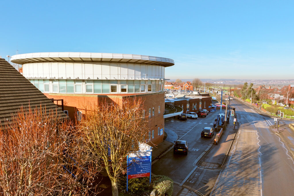 Photograph of Montagu Hospital using a drone. Angle is raised in the air level with the highest point looking out at the entrance. It is a sunny day with a clear blue sky. 