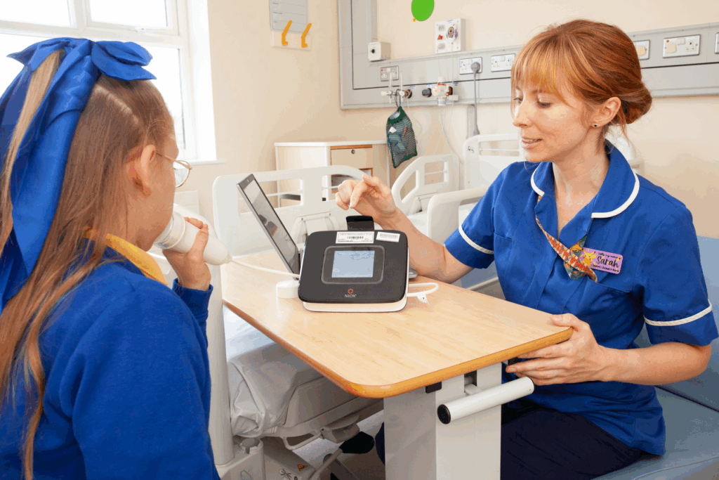 Photograph of a female nurse with ginger hair and blue unfirm sat at bedside table in a ward area. She is pointing at a digital tool on the table. A young girl sat opposite with a blue uniform and bow in her hair blows into a device connected to the device on the table. 