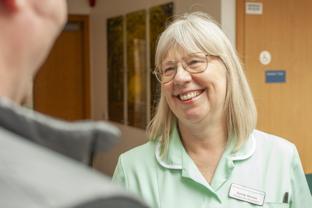Photograph of a female healthcare assistant with blonde hair, round glasses and a light green uniform smiling at a patient who is cropped and  blurred in the foreground.  