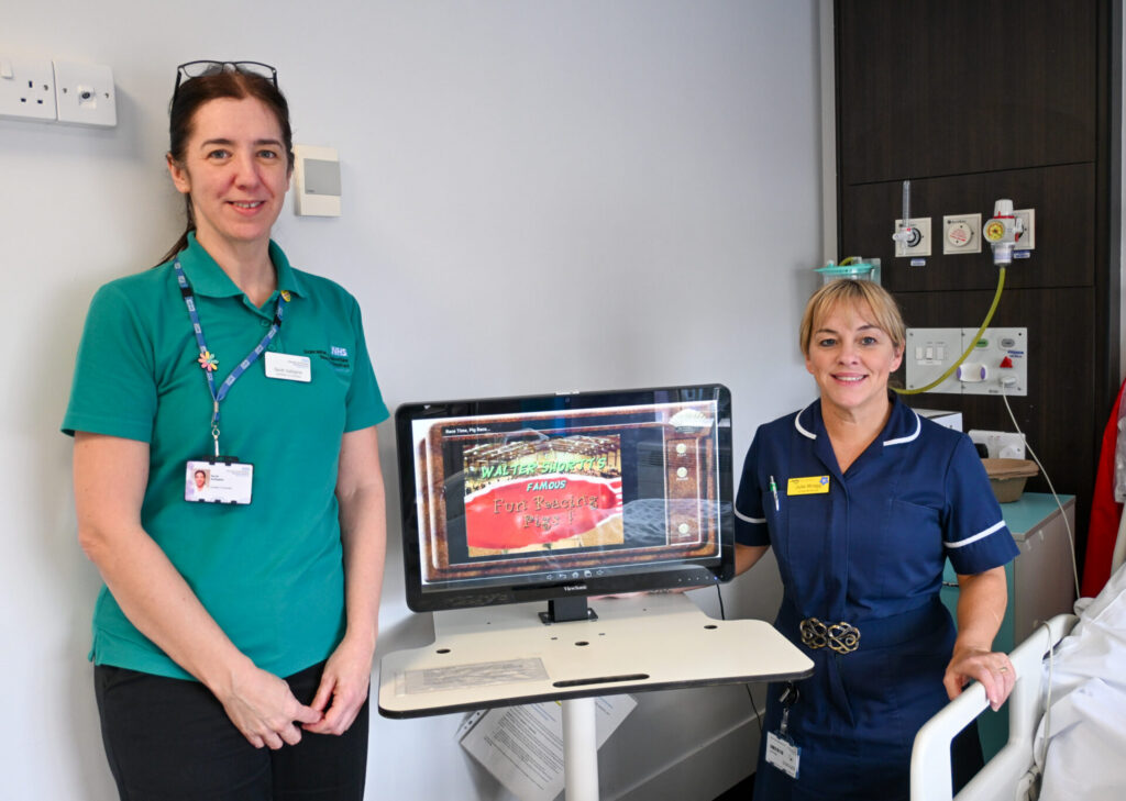 Julie and Sarah stand either side of RITA wearing their uniforms by a patient bedside. RITA is a large screen on a stand, and one of the activities you can interact with is on display. 