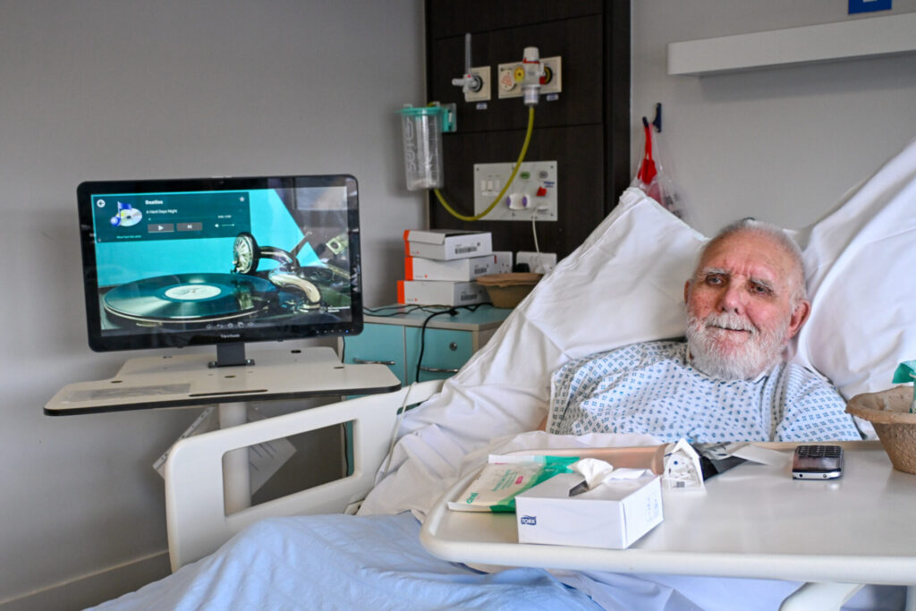 Patient Trevor, a older gentleman with white facial hair, is in a hospital bed looking at the camera in a hospital gown. By by the bedside the RITA screen is set up playing music. 