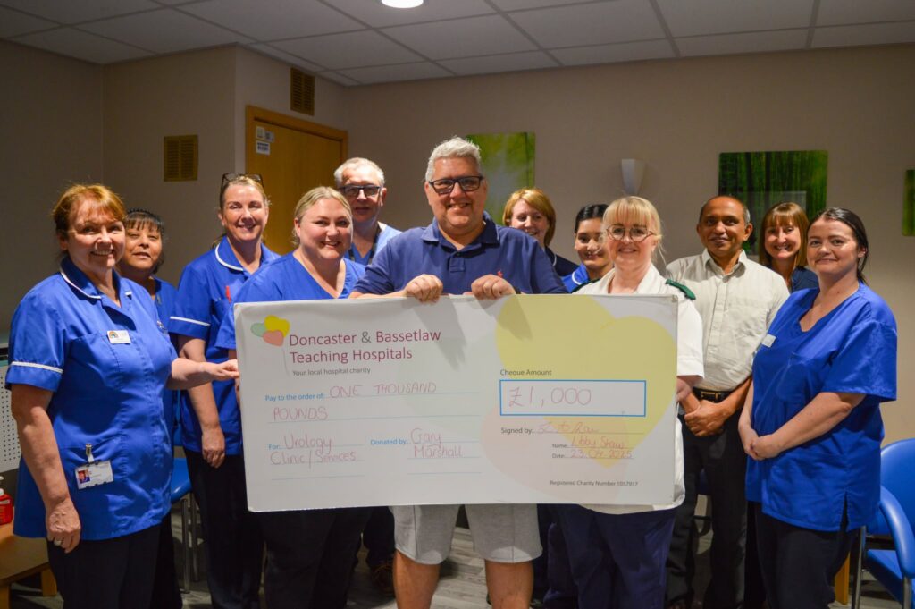 Gary, a white middle aged man, stands in the middle of a group of healthcare staff in blue uniforms. They are all smiling at the camera and Gary is holding a large cheque displaying £1,000