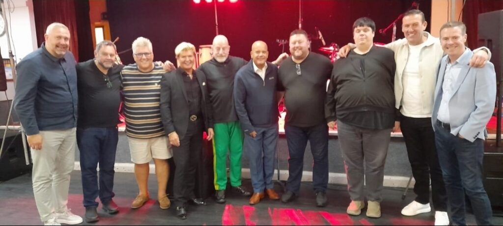 A group photo of nine men and one woman stood in front of a stage with red lighting.