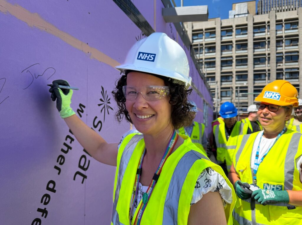 Zara is wearing a white hard hat and yellow high-vis jacket. Smiling at the camera, her gloved hand is signing a construction board