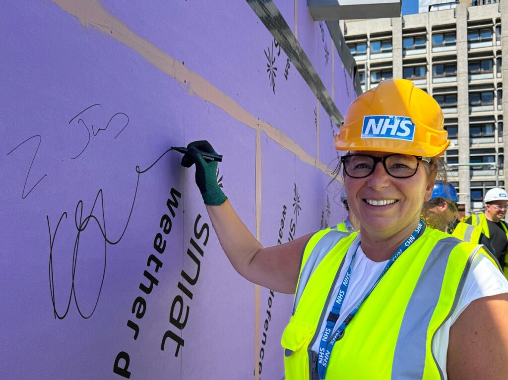 Kirsty is wearing a white hard hat and yellow high-vis jacket. Smiling at the camera, her gloved hand is signing a construction board
