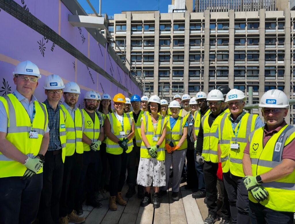 A group of people wearing white hard hats and yellow high-vis jackets stand in front of the construction outside the hospital.