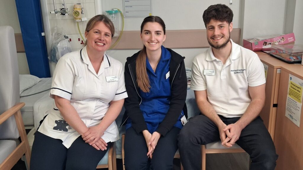 Three healthcare staff in blue and white uniforms sit on chairs in a ward area, smiling at the camera.