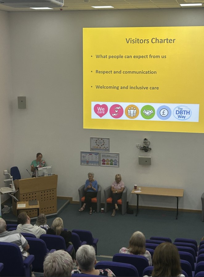 View of a person sat in lecture theatre seating looking at screen with a yellow background and black writing. Speaker stands to the left behind a lectern.