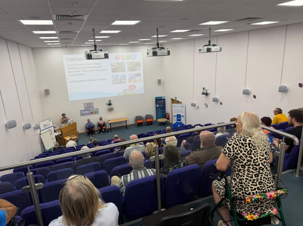 View from the top of the lecture theatre, looking down at patients sat in rows of blue seating looking at the screen and speaker at the front.