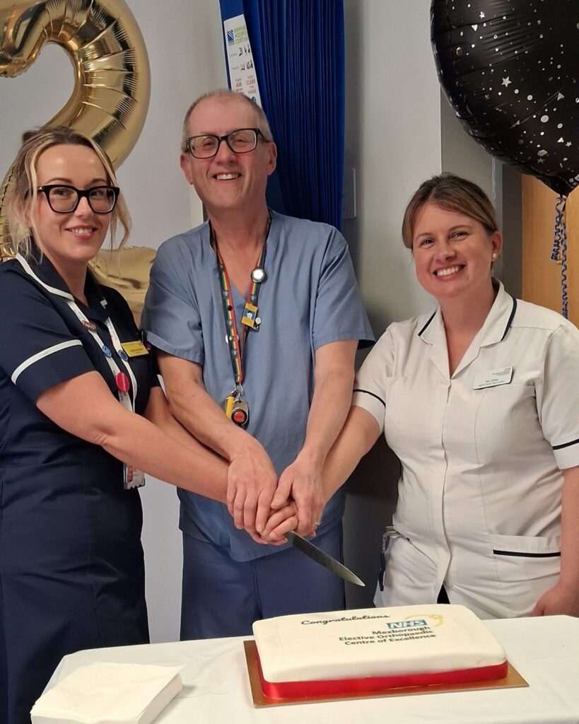 Three healthcare staff in uniforms and scrubs smile a the camera, each with a hand on a knife that's cutting a cake on the table in front of them. The icing on the cake reads congratulations.