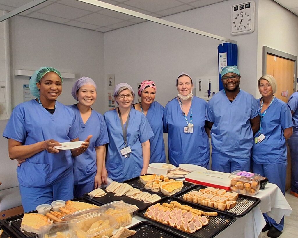 Group of 7 healthcare staff in blue scrubs are smiling at the camera in front of a table of picnic food, including sandwiches, sausage rolls and quiche.