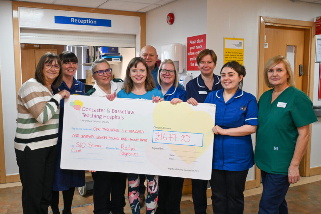 Rachel, a white woman with brown hair, stands in the centre of a group of people holding a large cheque that reads £1677.20. Around her are nurses in blue uniforms smiling at the camera.